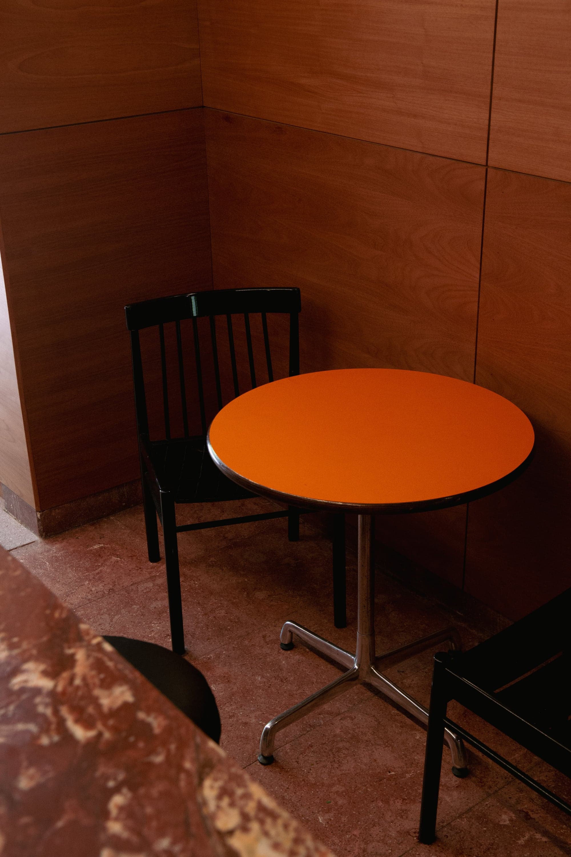 Una mesa redonda laqueada en naranja junto a una silla negra de madera, en un rincón de paredes de madera